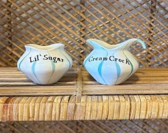 May include: Two ceramic sugar and cream containers. The sugar bowl is labeled "Lil' Sugar" and the cream pitcher is labeled "Cream Crock". Both are white with blue vertical stripes and a scalloped rim.