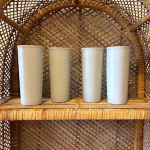 May include: Four tall, cylindrical, light-colored ceramic tumblers are displayed on a woven shelf. The tumblers are evenly spaced and have a smooth, matte finish. The shelf is part of a larger woven structure.