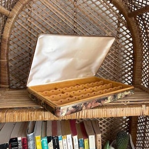 Peut inclure: Une boîte à bijoux rectangulaire avec un couvercle à charnière, présentant un extérieur à motif floral, une bordure dorée métallique et une doublure intérieure en satin crème. La base contient un insert en velours jaune moutarde.