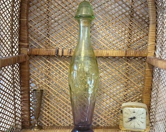 May include: A green and purple glass decanter with a textured surface sits on a woven wicker surface. The decanter has a wide base and a narrow neck with a green glass stopper.