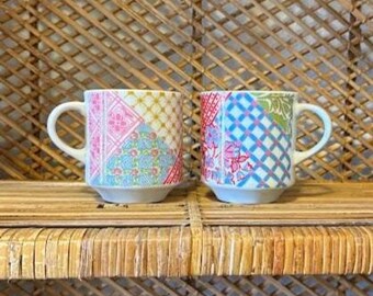 Może przedstawiać: Dwa ceramiczne kubki z białymi uchwytami i podstawami. Każdy kubek ma wzór patchwork z różowymi, niebieskimi i żółtymi wzorami kwiatowymi i geometrycznymi. Kubki są wystawione na plecionej półce.