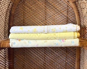 Peut inclure: Trois draps anciens roulés, dans les tons de jaune et de blanc, empilés sur une étagère en osier. Le drap supérieur présente un motif floral et une bordure en dentelle. Le drap du milieu est jaune pâle uni, et le drap du bas a un motif floral.