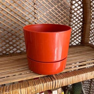 May include: A cylindrical orange plastic plant pot with a smooth, glossy finish. The pot features a distinct horizontal seam near the base, separating the main body from the lower section, creating a simple, functional design for holding plants.