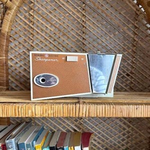 May include: A vintage General Electric electric pencil sharpener. The sharpener has a brown front panel with the word "Sharpener" printed on it. The sharpener has a silver top and a white border. The sharpener is sitting on a wooden shelf.