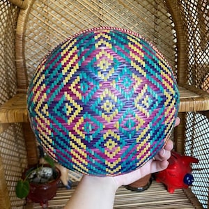 Könnte beinhalten: Ein runder, gewebter Korb mit einem lebendigen geometrischen Muster in Lila, Türkis, Gelb und Rosa. Der Korb wird in einer Hand gehalten und scheint aus Naturfasern hergestellt zu sein. Das Design weist Rautenformen auf.