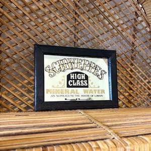May include: A framed vintage advertisement for Schweppes Mineral Water. The black framed sign features the Schweppes logo and the words "High Class Mineral Water" in gold lettering. The sign is displayed on a woven bamboo surface.