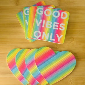May include: A collection of coasters in two shapes: rectangular and heart-shaped. The rectangular coasters display the text "GOOD VIBES ONLY" in white, set against a rainbow striped background. The heart-shaped coasters feature a similar rainbow striped design.