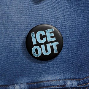 May include: A black button with the words "ICE OUT" in light blue, distressed font. The button is round and set against a blue denim background. The text is the focal point, with a cracked ice effect.