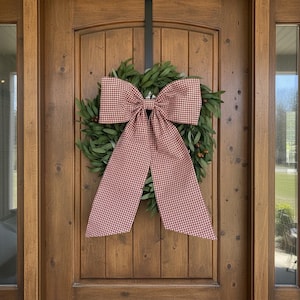 May include: A round wreath made of green leaves and small brown berries, featuring a large, prominent bow made of fabric with a red and white gingham check pattern. The bow has two long, wide tails that hang down over the center of the foliage.