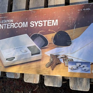 May include: A white box with the text "4-STATION INTERCOM SYSTEM" on the front. The box is open and shows a white intercom unit with a black dial and a black button. The box also includes a pair of sunglasses, a key chain, and a set of four smaller intercom units.