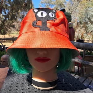 May include: Orange bucket hat with a black cat appliqué. The cat has large white eyes and a black outline.