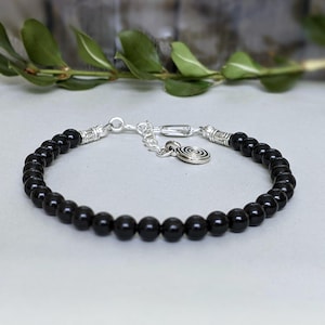 May include: A black beaded bracelet with silver-coloured metal accents and a spiral charm. The bracelet features small, round, black beads strung together with a silver chain and clasp. Green leaves are in the background.
