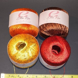 May include: Four spools of Ella Rae Sequinicity yarn in orange, brown, gold, and red. Each spool has a white label with the brand name and the words "Sequinicity adds sparkle to any project".