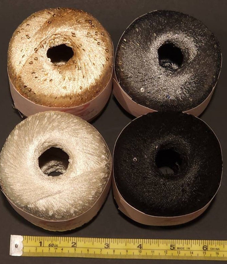 Puede incluir: Cuatro carretes de hilo decorativo en oro, plata, negro y gris oscuro. Cada carrete tiene forma de rosquilla con un agujero central y est&aacute; envuelto en una etiqueta de papel. El hilo parece tener una textura con lentejuelas o brillante.
