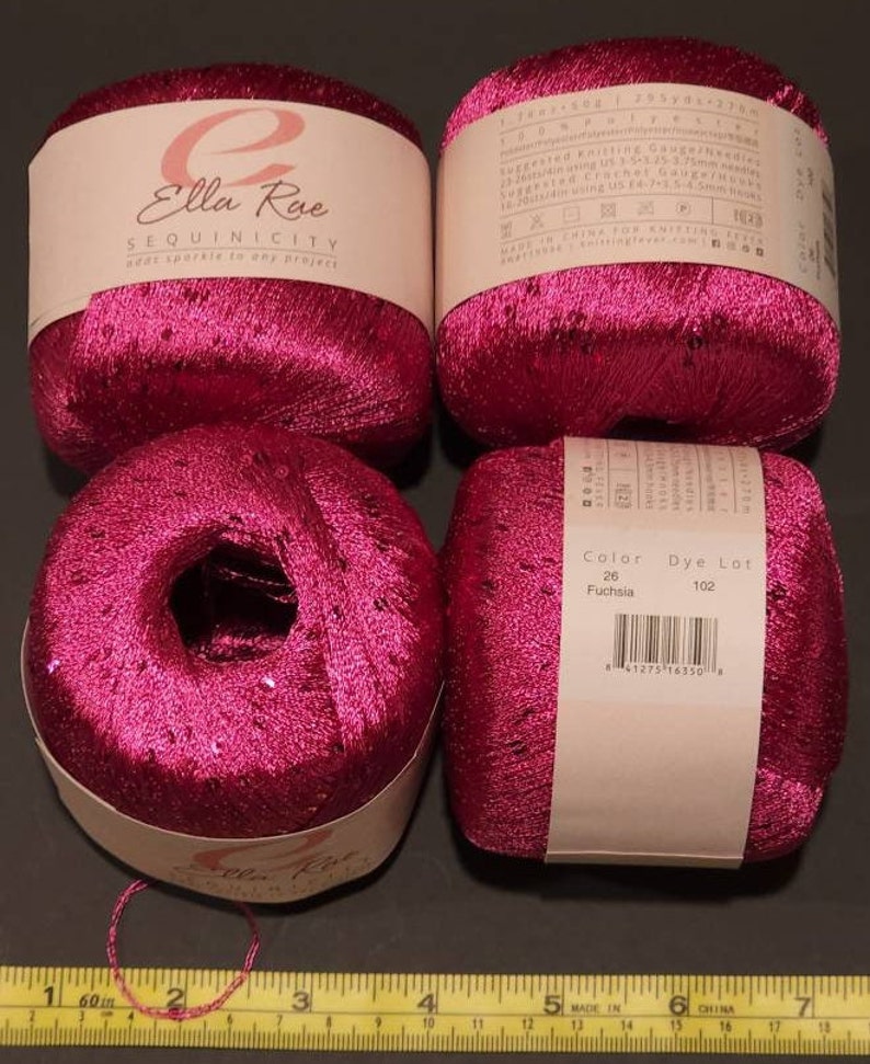 Pu&ograve; includere: Quattro rocchetti di filato rosa fucsia con paillettes. Il filato &egrave; etichettato "Ella Rue Sequinicity add sparkle to any project". L'etichetta include anche il numero di colore 26, il lotto di tintura 102 e un codice a barre.