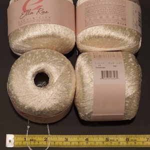 Puede incluir: Cuatro madejas de hilo de lentejuelas blanquecino con etiquetas rosas. Las etiquetas dicen "Ella Rae SEQUINICITY" y "Color Dye Lot". Una cinta m&eacute;trica muestra 152 cm. El hilo es para proyectos de tejido y ganchillo.