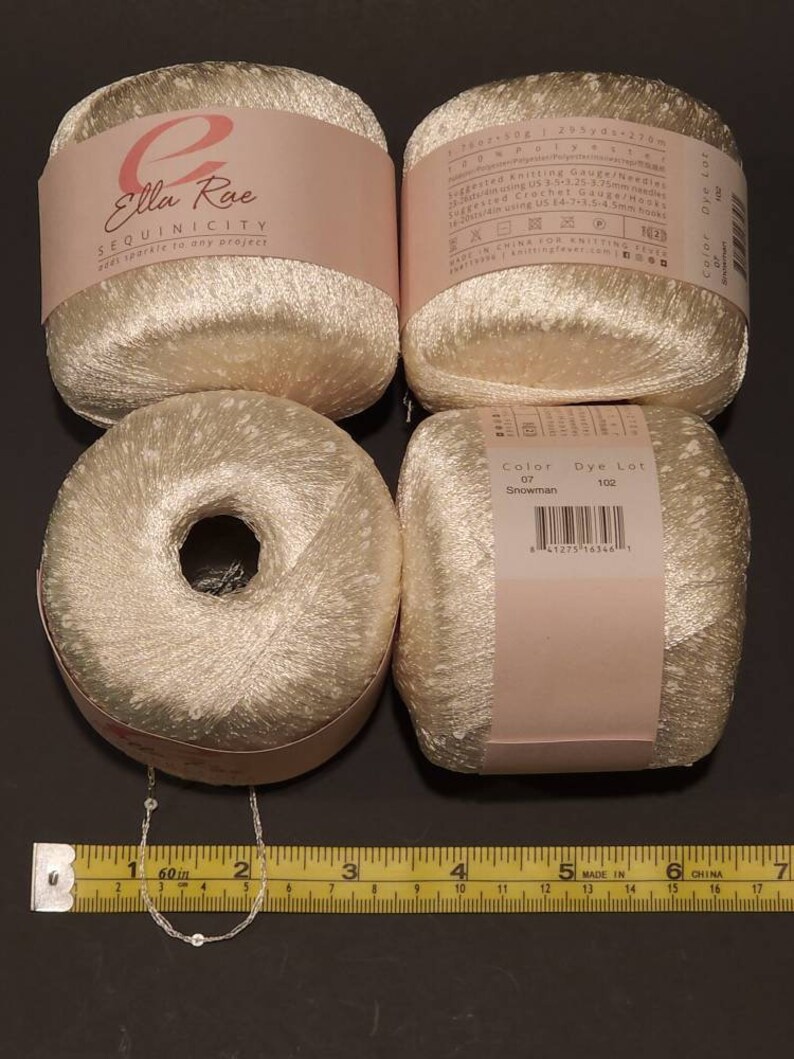 Puede incluir: Cuatro madejas de hilo de lentejuelas blanquecino con etiquetas rosas. Las etiquetas dicen "Ella Rae SEQUINICITY" y "Color Dye Lot". Una cinta m&eacute;trica muestra 152 cm. El hilo es para proyectos de tejido y ganchillo.