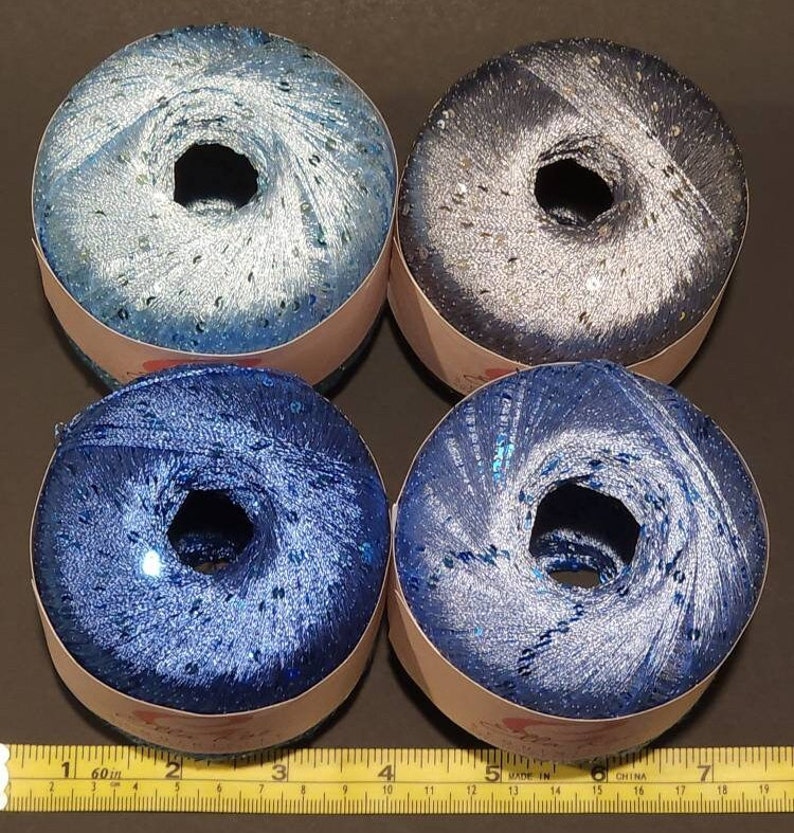 May include: Four spools of blue and silver yarn with sequins. The yarn is wound around a cardboard spool with a white label.