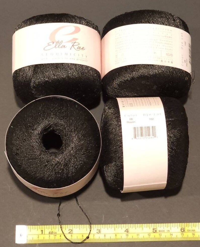 Puede incluir: Cuatro ovillos de hilo negro con etiquetas rosas y blancas. Las etiquetas dicen "Ella Rae Sequinsicity". Una cinta m&eacute;trica amarilla muestra 152 cm. El hilo es de color oscuro y brillante, y los ovillos son redondos.