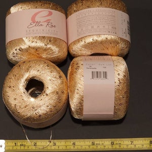 Puede incluir: Cuatro bobinas de hilo de lentejuelas doradas dispuestas sobre una superficie oscura. El hilo est&aacute; etiquetado como "Ella Rae SEQUINICITY" y "Sandcastle". Una cinta m&eacute;trica est&aacute; en la parte inferior. Cada bobina tiene unos 5 cm de di&aacute;metro.