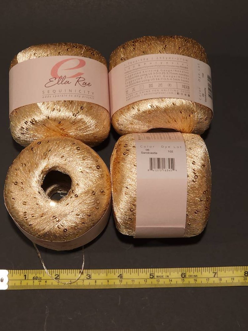 Puede incluir: Cuatro bobinas de hilo de lentejuelas doradas dispuestas sobre una superficie oscura. El hilo est&aacute; etiquetado como "Ella Rae SEQUINICITY" y "Sandcastle". Una cinta m&eacute;trica est&aacute; en la parte inferior. Cada bobina tiene unos 5 cm de di&aacute;metro.