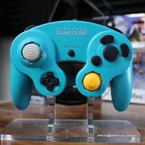 May include: A teal Nintendo GameCube controller with black buttons and a yellow button on the bottom right. The controller is on a clear plastic stand.