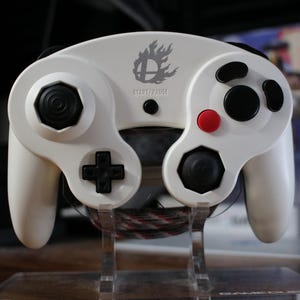 May include: A white video game controller with black and red buttons, a black directional pad, and a logo with the text "START/PAUSE". The controller is designed for gaming.