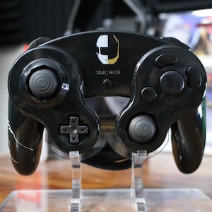 May include: A black video game controller featuring a white and gold split-face graphic in the center above the START/PAUSE button. The controller has a circular analog stick on the left, a D-pad below it, and a cluster of four buttons on the right.
