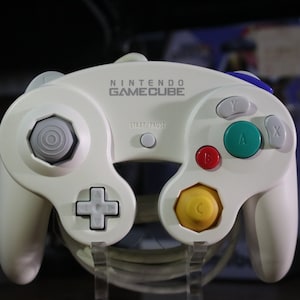 May include: Off-white Nintendo GameCube controller with the GameCube logo. Features a gray directional pad, a yellow octagon-shaped button, and colored buttons: red, green, and blue. The Start/Pause button is centered above the analog stick.