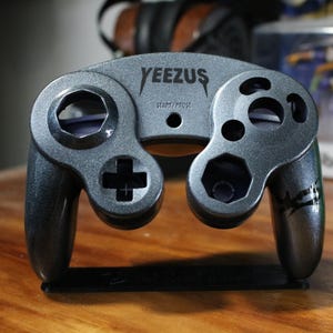 May include: A black and silver GameCube controller with the word "YEEZUS" printed on the front. The controller has a black and silver design with a black and white graphic on the side.