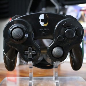 May include: A black GameCube controller with a white and gold Daft Punk helmet design on the front. The controller is on a clear plastic stand.