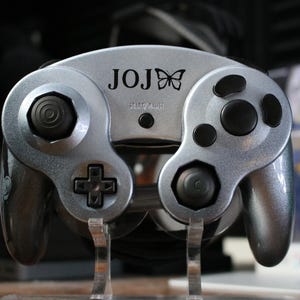 May include: A silver GameCube controller with a black butterfly logo and the text "JOJ" printed on the front. The controller has a black and silver design with a textured surface.