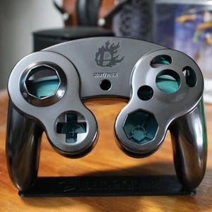 May include: A black GameCube controller with a blue and silver design. The controller has a "Super Smash Bros." logo on the top and the words "Start/Pause" below it.