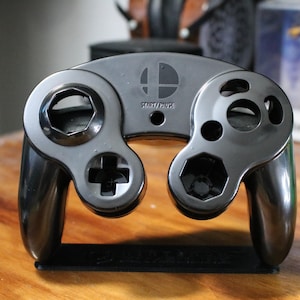 May include: A black video game controller with a glossy finish, featuring a directional pad, analog stick, and various buttons. The controller has the text "START/PAUSE" and a logo on the top. It is resting on a black stand.