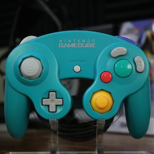 May include: Teal Nintendo GameCube controller with gray, red, green, and yellow buttons. The controller has the Nintendo GameCube logo and Start/Pause text. It is displayed on a clear stand.