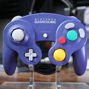 May include: A purple Nintendo GameCube controller with various colored buttons, including red, green, and yellow. The controller has the words "NINTENDO GAMECUBE" printed on the top. The controller is displayed on a clear stand.