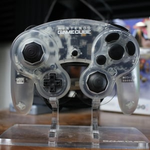 May include: A clear Nintendo GameCube controller with black buttons and joysticks. The controller is on a clear stand, with the GameCube logo visible. The controller has a transparent design, showcasing its internal components.