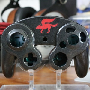 May include: A black metallic video game controller shell featuring a red fox silhouette logo centered above the joystick openings. The controller housing has cutouts for buttons, a D-pad, and joysticks, with a glossy finish and a dark interior.