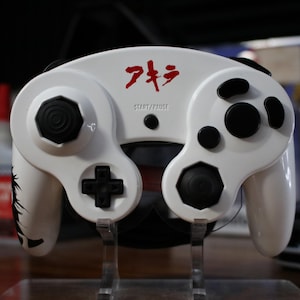 May include: White GameCube controller with black buttons and a red "アタック" logo on the top. The controller is on a clear stand.