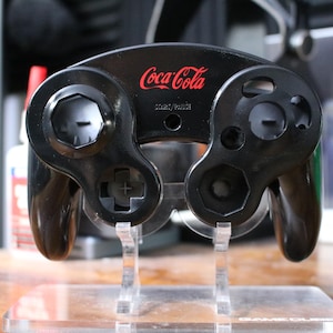 May include: Black GameCube controller with the Coca-Cola logo in red. The controller has various buttons and a directional pad. It is displayed on a clear acrylic stand.