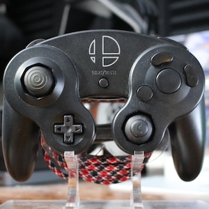May include: Black GameCube controller with the Super Smash Bros. logo and the text "SILENT/MOUSE" in white. The controller has a D-pad, analog stick, and buttons. A red and black braided cable is visible.
