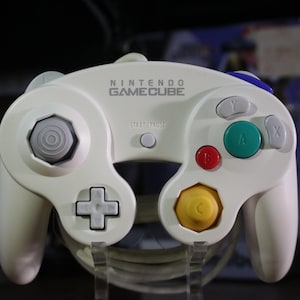 May include: A white Nintendo GameCube controller featuring a gray analog stick, a yellow C-stick, a green A button, a red B button, gray X and Y buttons, a gray D-pad, and a central Start/Pause button. The Nintendo GameCube logo is printed in gray.