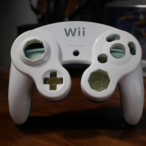 May include: A white Nintendo Wii GameCube controller with blue accents. The controller has a  "Wii" logo on the top and a "START/PAUSE" label below it.