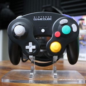 May include: Black Nintendo GameCube controller with colorful buttons, including red, yellow, green, and blue. The controller is displayed on a clear acrylic stand. The text "NINTENDO GAMECUBE" is printed on the top.