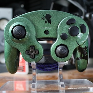 May include: A metallic green video game controller with black buttons, joysticks, and a directional pad. The controller features a black NERV logo and text on the front face, resting on a clear acrylic stand with a visible purple cable.