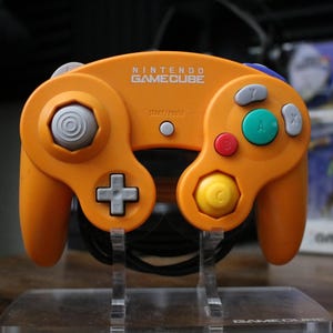 May include: Orange Nintendo GameCube controller with gray, red, yellow, and green buttons. The controller has a directional pad, analog stick, and a start button. The text "NINTENDO GAMECUBE" is printed on the top.