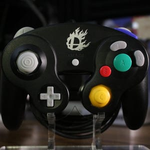 May include: A black video game controller featuring a white Super Smash Bros flame logo in the center. The device includes a large green A button, a red B button, gray X and Y buttons, a white directional pad, a white analog stick, and a yellow C-stick.
