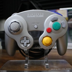 May include: Silver Nintendo GameCube controller with a variety of colored buttons, including red, green, and yellow. The controller has a directional pad and analog sticks. The words "NINTENDO GAMECUBE" are printed on the top.