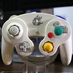May include: A white video game controller featuring a central Smash Bros logo and the text START/PAUSE. It includes a large gray analog stick, a green A button, a red B button, gray X and Y buttons, a yellow C-stick, and a white directional pad.
