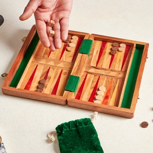 May include: A wooden backgammon set with red and green accents, dice, and game pieces. A green velvet drawstring bag and a wooden mustache and glasses stand are also present. The game is open, ready to play.